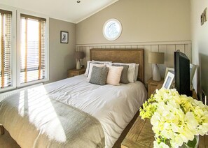 A bedroom with a bed, two bedside tables, a television, a window with blinds, and a circular skylight.