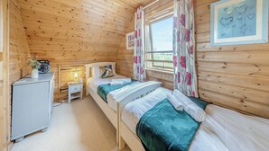 A cozy wooden cabin room with two beds, a small desk, a radio, and a window with floral curtains.