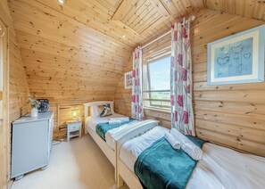 Ein gemütliches Holzhütten-Zimmer mit zwei Betten, einem kleinen Schreibtisch, einem Radio und einem Fenster mit blumigen Vorhängen.