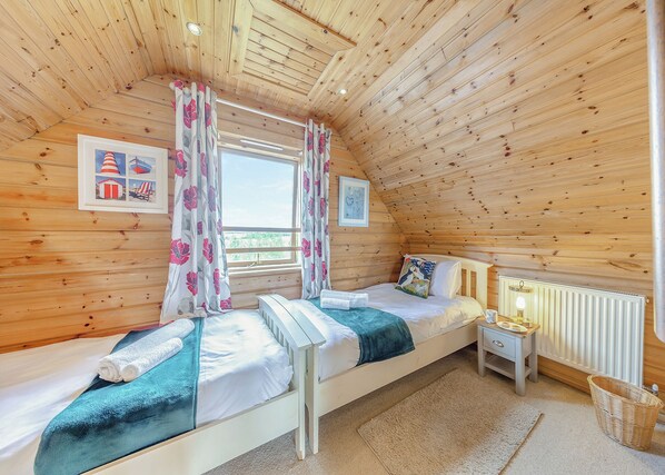 A cozy attic bedroom with wooden walls, a window with curtains, a bed with pillows, a bedside table with a lamp, and a radiator.