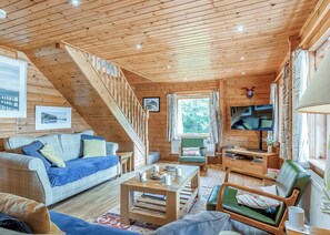 Ein gemütliches Blockhauss-Interieur mit einer Holztreppe, einem Sofa, einem Sessel, einem Couchtisch und einem Fernseher.