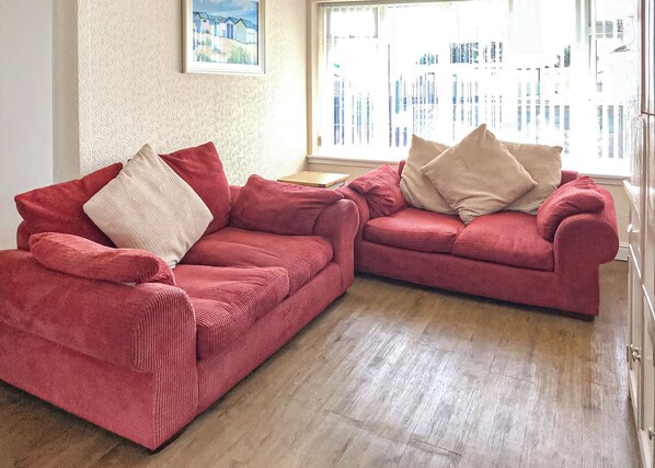 Two red sofas with cushions, a wooden floor, a patterned wall, and a window with a view of buildings. - Comfortable bungalow with one double and one twin. (Chapel St Leonards)
