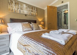 A hotel room with a large bed, a wooden headboard, a painting on the wall, a bathroom visible through an open door, and a nightstand with a lamp.