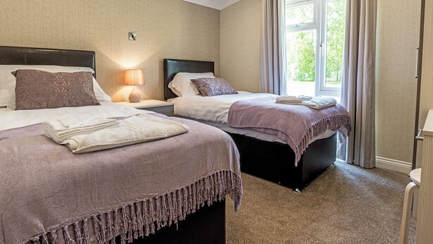 A hotel room with two beds, a window with curtains, a nightstand with a lamp, and a wall-mounted light.