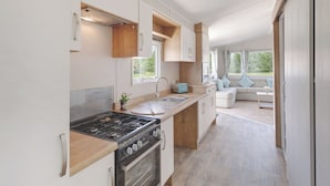 A modern RV kitchen with a stove, oven, and sink, leading to a living area with a sofa and windows.