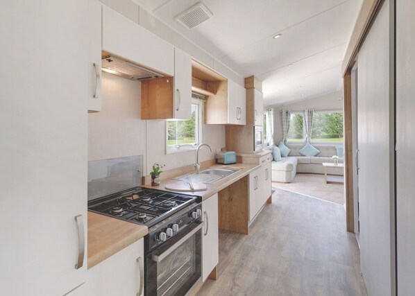A modern RV kitchen with a stove, oven, and sink, leading to a living area with a sofa and windows.