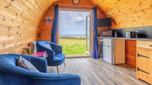 L’intérieur d’un chalet accueillant, avec un fauteuil bleu, une petite table sur laquelle repose un livre, un coin cuisine et une grande fenêtre donnant sur le paysage extérieur.