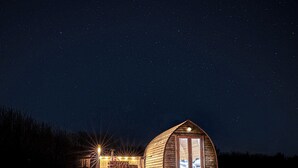 Une cabane en bois dotée d’une grande fenêtre et d’un toit en pente, entourée d’un champ verdoyant sous un ciel étoilé.