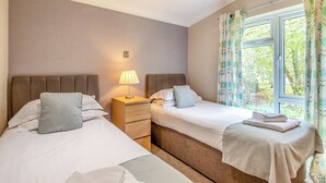 A hotel room with two beds, a bedside table with a lamp, a window with curtains, and a view of greenery outside. - 2 bedroom accommodation in Woodhall Spa (Woodhall Spa)