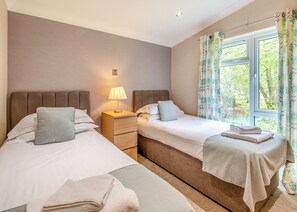 A hotel room with two beds, a bedside table with a lamp, a window with curtains, and a view of greenery outside. - 2 bedroom accommodation in Woodhall Spa (Woodhall Spa)