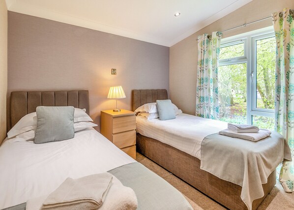 A hotel room with two beds, a bedside table with a lamp, a window with curtains, and a view of greenery outside.