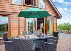 A wooden house with a glass door labeled 'CADNANT,' a table with chairs, a green umbrella, and a vase with flowers. - Spacious, light, all wood luxury lodge in tranquil location at head of lake. (Anglesey)