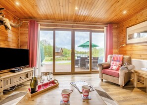 L’intérieur d’un chalet en bois comprend une télévision, une table à manger avec des verres à vin, un canapé recouvert d’une couverture à carreaux et une grande porte-fenêtre coulissante donnant sur une terrasse extérieure.