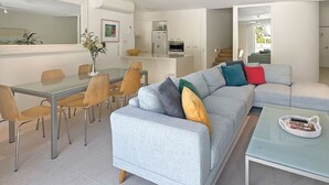 A modern living room with a grey sofa, a glass coffee table, and a dining area with a glass table and wooden chairs. - Penthouse 23 - The Cove (Noosa)