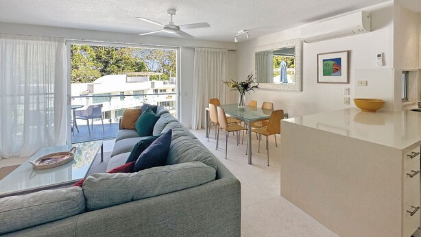 A modern living room with a grey sofa, a glass coffee table, and a dining area with a glass table and chairs. - Penthouse 23 - The Cove (Noosa)