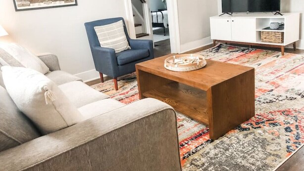 Una sala de estar con sofá, una mesita de centro, un sillón azul, un televisor sobre un mueble blanco y una alfombra estampada.