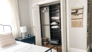 Un dormitorio con cama, mesita de noche, lámpara, armario abierto y un adorno de macramé en la pared.