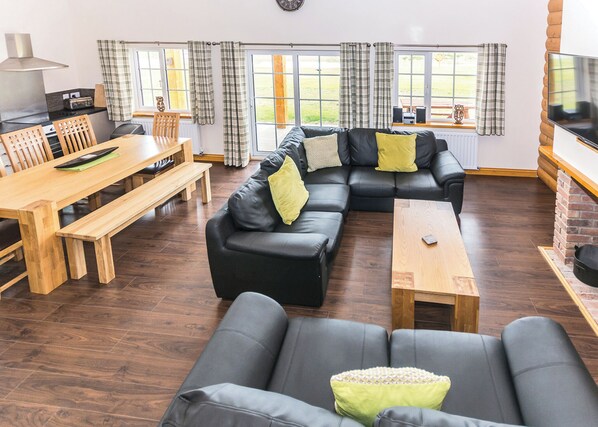 A modern living room with a sectional sofa, a wooden dining table, and a TV mounted on a brick wall.