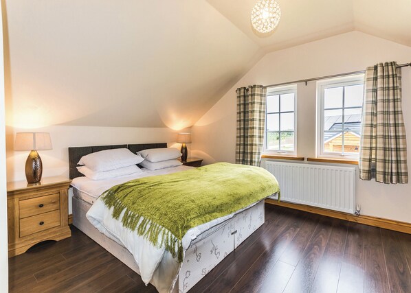 Un dormitorio con cama, dos mesitas de noche, lámpara, ventana con cortinas y radiador.