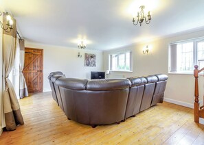 A spacious living room with a large leather sofa, wooden flooring, a chandelier, and a view through a window. - Three bedroomed holiday cottage. (Swinethorpe, Nr Lincoln)