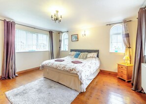 A bedroom with a large bed, a chandelier, a nightstand, and a window with curtains. - Three bedroomed holiday cottage. (Swinethorpe, Nr Lincoln)