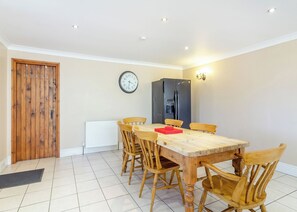 Een eetgedeelte met een houten tafel, zes stoelen, een klok, een koelkast en inbouwspots.