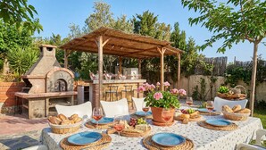 Un espacio de comedor al aire libre con un horno de ladrillo, una zona de estar cubierta y una mesa dispuesta para una comida.