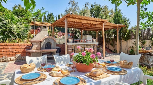 An outdoor dining area with a brick oven, a covered seating area, and a table set for a meal.