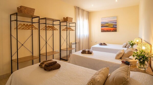 Habitación de hotel con dos camas, un armario con ropa colgada, un cuadro en la pared y una mesita de noche con una lámpara y flores.