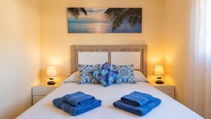 Una cama bien hecha con toallas azules, almohadones estampados y un cuadro de atardecer arriba.