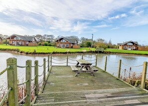 Une jetée en bois avec une table de pique-nique, une rivière paisible et deux cabanes en bois en arrière-plan.