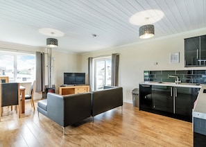A modern living room with a flat-screen TV, a sofa, a dining area, and a kitchen with black cabinets and a stainless steel sink.