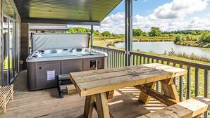 Un terrazzo in legno con vasca idromassaggio, tavolo da picnic e panchine con vista su uno specchio d'acqua.