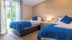 A hotel room with two beds, a nightstand, a lamp, and a window with curtains. - Lakeside 3 Pet Friendly (Woodhall Spa)