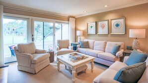 Una sala de estar con un sofá color beige, dos sillones, una mesa de centro y una mesita auxiliar con una lámpara. Se observan obras de arte enmarcadas en la pared y vistas al exterior a través de amplias puertas correderas.