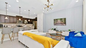 Ein modernes Wohnzimmer mit einem weißen Sofa, einem blauen Sessel und einer gelben Decke. Es gibt eine Küchenzeile mit dunklen Schränken und einer weißen Arbeitsplatte. An der Wand hängt ein Bild von einer Waldstraße.