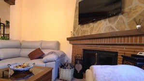 A living room with a stone fireplace, a flat-screen TV mounted above it, a beige sofa, and a coffee table with a bowl of fruit. - Casa Rural El Ojuelo (VTAR) (El Ojuelo)