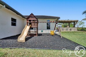 A playground with a slide and swings, a house with a sloped roof, and a stone wall in the background. - 8-BR with Pool & Mini-golf & Soccer (Davie)
