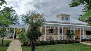 A two-story building with a white exterior and a yellow roof, surrounded by lush greenery and a stone pathway. - La caz à Anne, Villa de Charme (Saint Paul)
