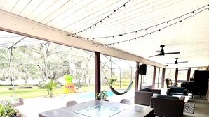 Eine überdachte Terrasse im Freien mit einem Glastisch und Stühlen, umgeben von Sitzgelegenheiten und einem Pool.