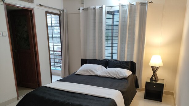 A bedroom with a bed, a nightstand with a lamp, a window with curtains, and a door. - Nicely 3 bedrooms apartment at Mohammadpur (Dhaka)