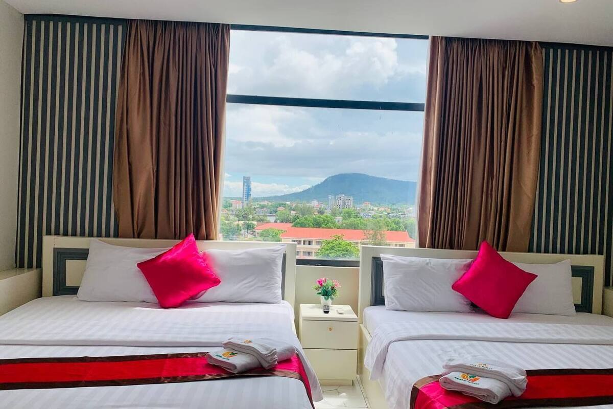 A hotel room with two beds, each with white bedding and a red accent. There is a window with curtains, and a view of a cityscape and mountains in the background.