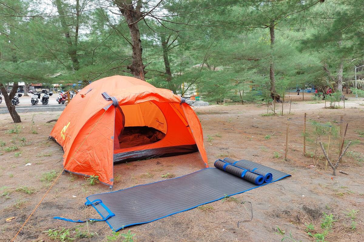Une tente orange est installée dans une zone boisée, avec un tapis et des tapis enroulés à proximité.