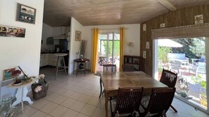 Uma área de refeições com mesa e cadeiras de madeira, cozinha ao fundo e vista para o exterior por uma grande janela.