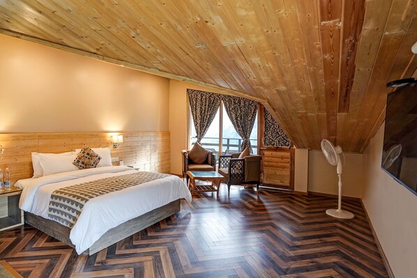 Une pièce chaleureuse avec un plafond en bois, un grand lit avec une literie à motifs, une petite table, deux chaises et un ventilateur.
