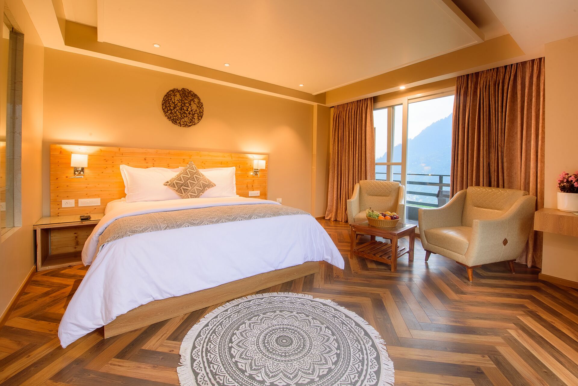 A hotel room with a large bed, two armchairs, a small table with fruit, and a view of mountains through the window.