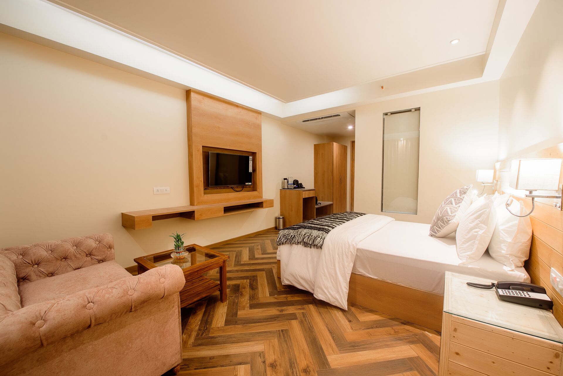 A hotel room with a bed, a TV mounted on the wall, a wooden nightstand, a small table with a plant, and a mirror.
