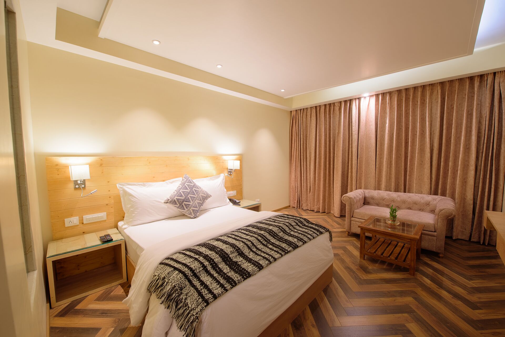 A hotel room with a large bed, a sitting area with a sofa, a wooden nightstand, and a glass coffee table.