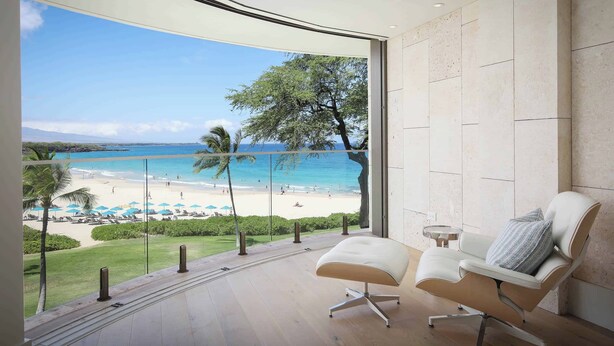 A modern room with a curved ceiling, a white chair with a pillow, and a view of a beach with palm trees and umbrellas. - Hapuna Beach Residences A34 Two Bedroom (Kamuela)