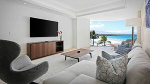 A modern living room with a flat-screen TV, a wooden cabinet, a grey armchair, a patterned sofa, and a view of a beach. - Hapuna Beach Residences A34 Two Bedroom (Kamuela)